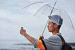 Asian travel man taking photo using smartphone camera photographing beautiful seaside holding transparent umbrella on rainy day
