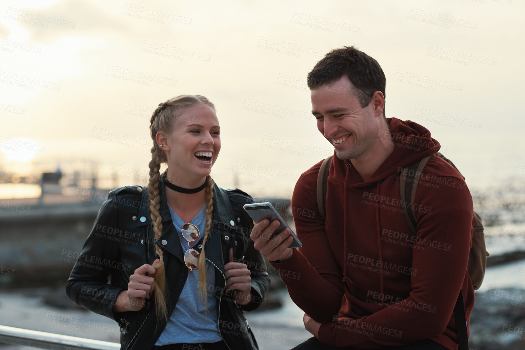 Buy stock photo Phone, friends and laugh by sea with social media, funny joke and text message with meme on vacation. Happy people, mobile and students at beach for humor, post or gossip at sunset on holiday break
