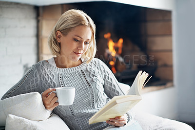 Buy stock photo A beautiful young woman reading a book and enjoying a warm beverage near a fireplace