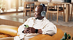 Music, phone and thinking with black man on sofa in home living room for me time or wellness. Headphones, relax and thoughtful with happy person in apartment for audio, song or sound streaming