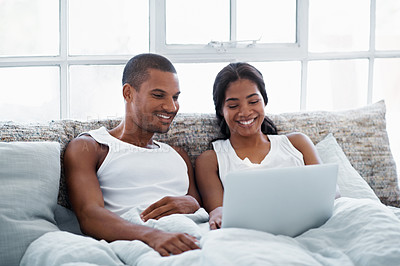 Buy stock photo Shot of a smiling young couple lying in bed using a laptop