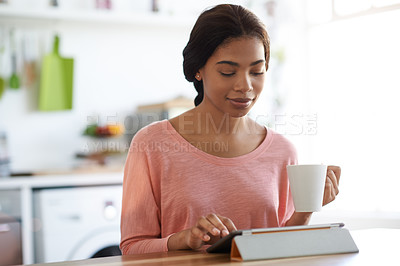 Buy stock photo Shot of a young woman drinking a coffee and using her digital tablet in the kitchen