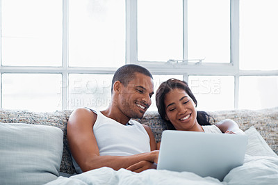 Buy stock photo Shot of a smiling young couple lying in bed using a laptop