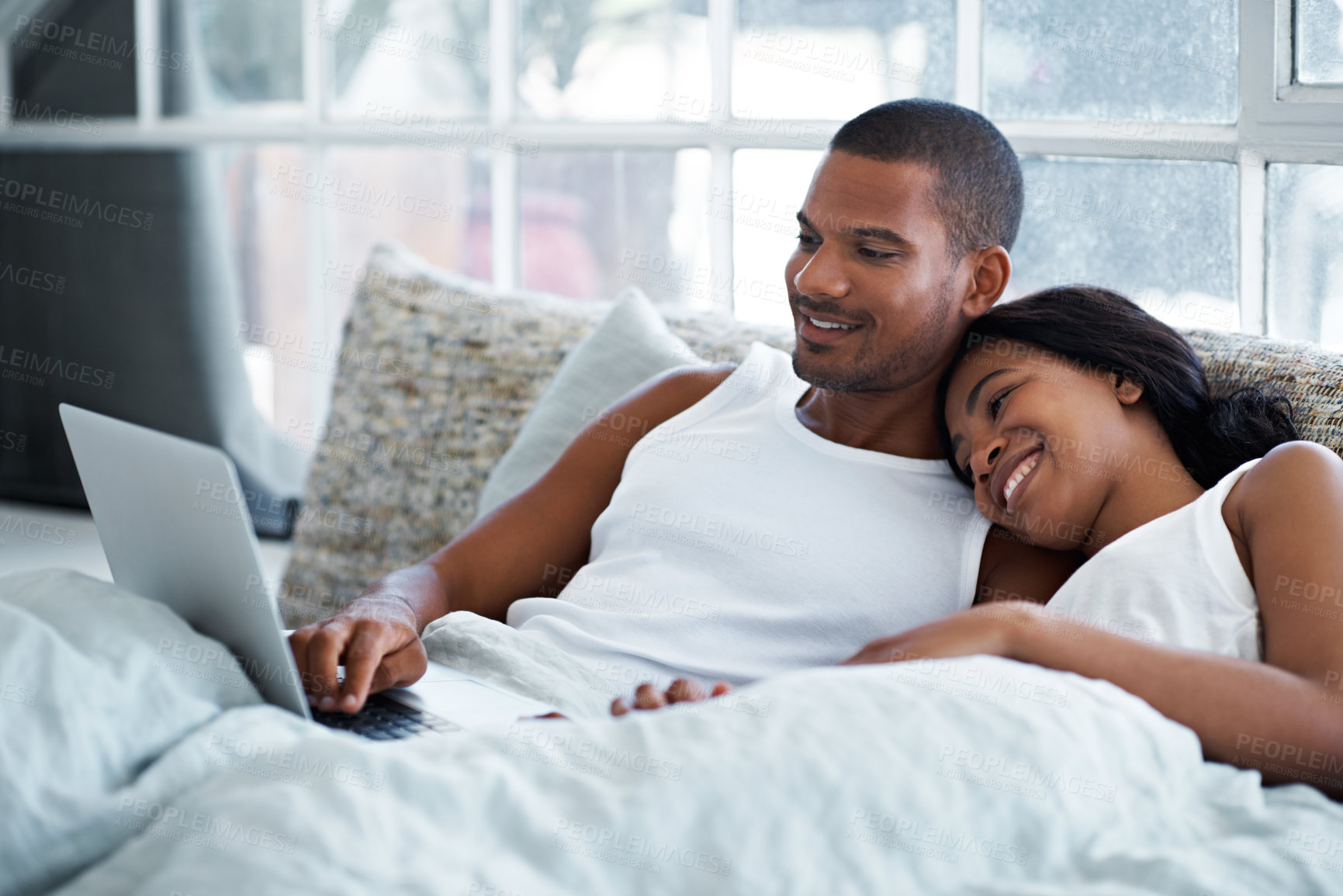 Buy stock photo Shot of a smiling young couple lying in bed using a laptop