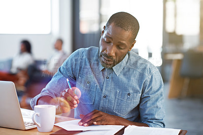 Buy stock photo Shot of a man taking notes and working on a laptop in an office with colleagues in the background