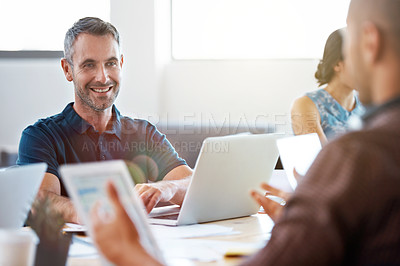 Buy stock photo Portrait of a smiling man sitting in an office working on a laptop surrounded by colleagues