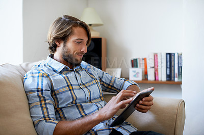 Buy stock photo Shot of a young man using a digital tablet while sitting on a sofa at home