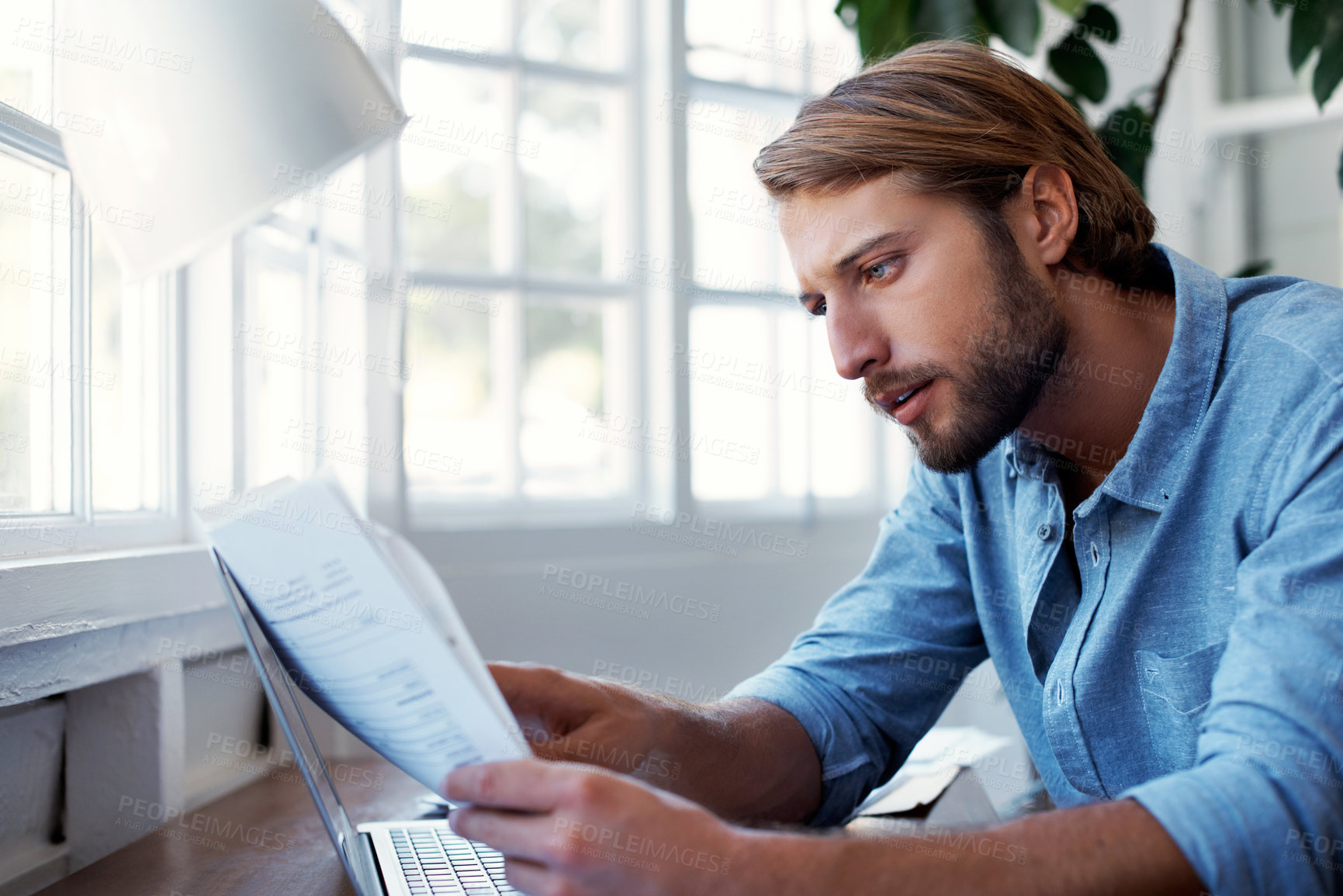 Buy stock photo Shot of a young man reading papers while working on a laptop at home