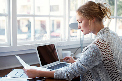 Buy stock photo Shot of a young woman reading papers while working on a laptop at home