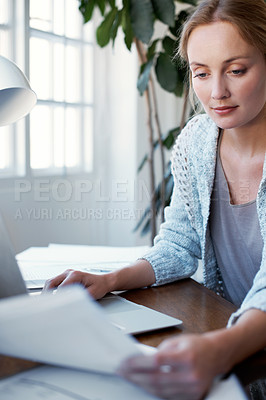 Buy stock photo Shot of a young woman reading papers while working on a laptop at home