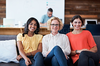 Buy stock photo Portrait of three beautiful coworkers sitting together in the office with colleagues blurred in the background