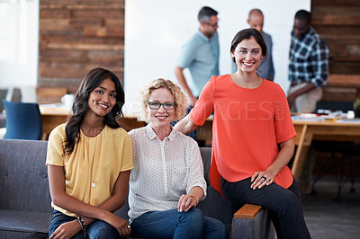 Buy stock photo Portrait of three beautiful coworkers sitting together in the office with colleagues blurred in the background