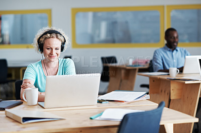 Buy stock photo Shot of a woman listening to music while working on her laptop with her colleague blurred in the background