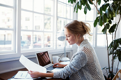 Buy stock photo Shot of a young woman reading papers while working on a laptop at home