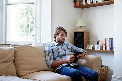Buy stock photo Shot of a young man using a digital tablet while sitting on the sofa at home