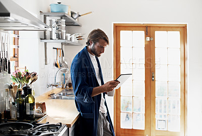 Buy stock photo Shot of a young man using his tablet while having coffee in the kitchen
