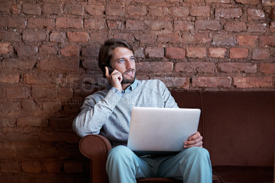Buy stock photo Shot of a young man talking on his cellphone and using a laptop while sitting at home