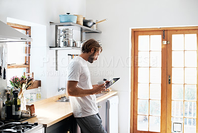 Buy stock photo Shot of a young man using his tablet while having coffee in the kitchen