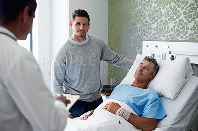 Buy stock photo Shot of a doctor talking to a man lying in a hospital bed while his adult son look on