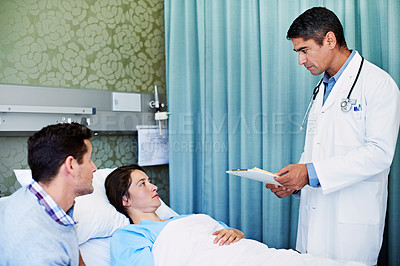 Buy stock photo Shot of a doctor talking to a woman lying in a hospital bed while her husband looks on