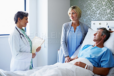 Buy stock photo Shot of a doctor talking to a man lying in a hospital bed with his wife beside him