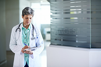 Buy stock photo Shot of a mature female doctor reading a paperwork while standing in a hospital corridor