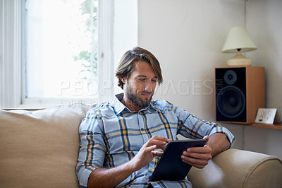 Buy stock photo Shot of a young man using a digital tablet while sitting on a sofa at home