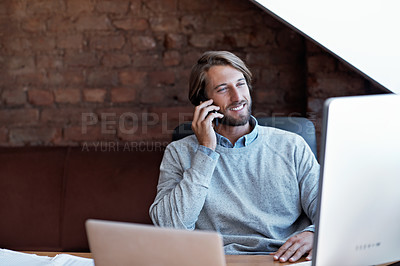 Buy stock photo Shot of a young man talking on the phone while working on a computer at home