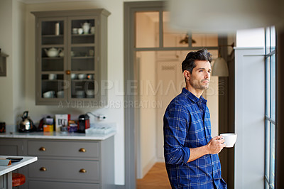 Buy stock photo Shot of a young man drinking a coffee while looking through his kitchen window