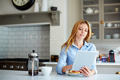 Buy stock photo Shot of a young woman using her digital tablet while sitting in her kitchen at home