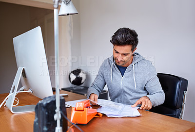 Buy stock photo Shot of a young man sitting at a desk in his home office reading documents and working on a computer