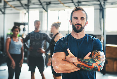Buy stock photo Cropped portrait of a handsome young male athlete standing with his arms folded in the gym with other gym-goers in the background