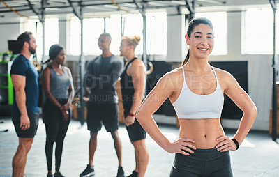 Buy stock photo Cropped portrait of an attractive young female athlete standing with her hands on her hips in the gym with other gym-goers in the background