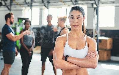 Buy stock photo Cropped portrait of an attractive young female athlete standing with her arms folded in the gym with other gym-goers in the background