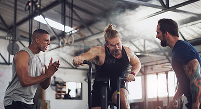 Buy stock photo Shot of a young man working out on an exercise bike while his friends encourage him in a gym
