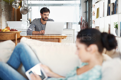 Buy stock photo Shot of a handsome young man using a laptop while his girlfriend is relaxing on the sofa