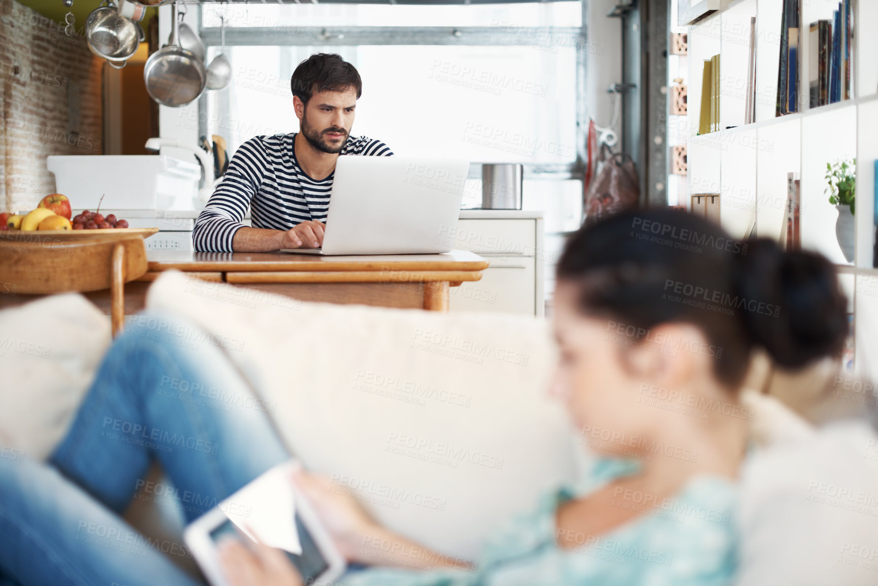 Buy stock photo Shot of a handsome young man using a laptop while his girlfriend is relaxing on the sofa