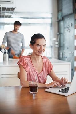Buy stock photo Shot of a beautiful young woman using a laptop while her husband is preparing food in the background
