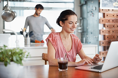 Buy stock photo Shot of a beautiful young woman using a laptop while her husband is preparing food in the background