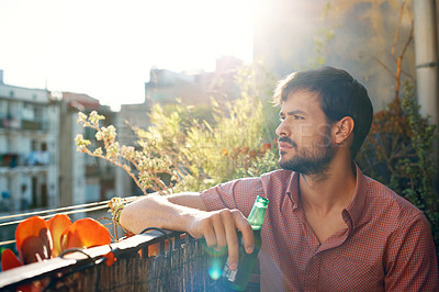 Buy stock photo Shot of a handsome young man holding a beer and looking thoughtful while sitting outside on a balcony