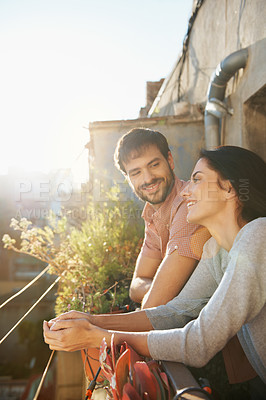 Buy stock photo Shot of a beautiful young couple standing next to each other on a balcony overlooking an urban setting