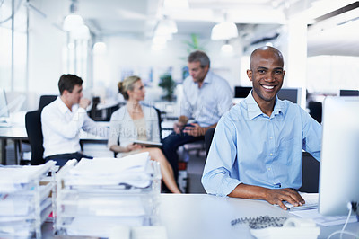 Buy stock photo Portrait of a smiling office worker sitting at his workstation with colleagues in the background
