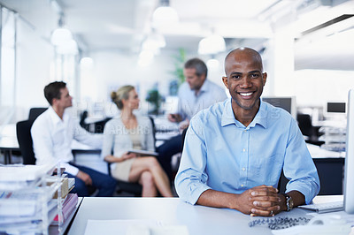 Buy stock photo Portrait of a smiling office worker sitting at his workstation with colleagues in the background