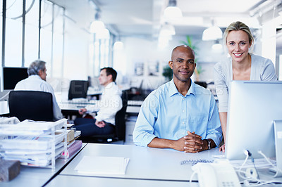 Buy stock photo Portrait of two office workers talking over a desktop computer in an office iwth colleagues in the background