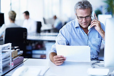 Buy stock photo Shot of a mature businessman talking on a phone and reading a document while sitting at his desk in an office