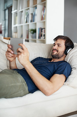 Buy stock photo A handsome young man listening to music on his tablet while lying on the sofa