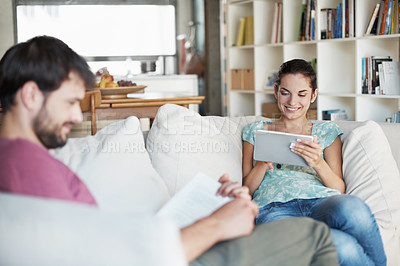 Buy stock photo A beautiful young woman using her tablet while her husband reads a book
