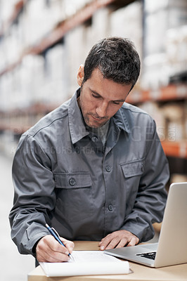 Buy stock photo A young man using his laptop and taking notes while working in a warehouse