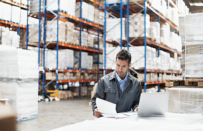 Buy stock photo A young man using his laptop and looking over his notes while working in a warehouse
