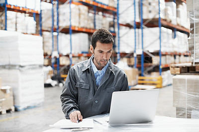 Buy stock photo A young man using his laptop and checking his notes while working in a warehouse